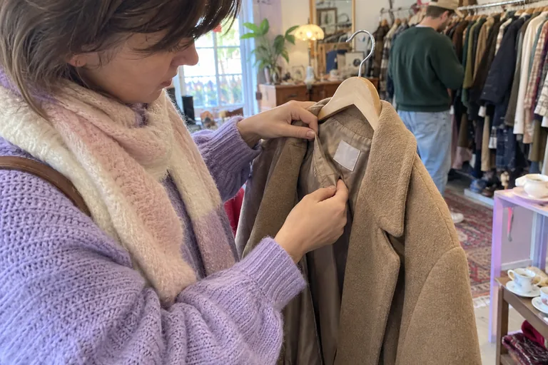 Des mains examinent les coutures et l’étiquette de composition à l’intérieur d’un manteau en laine suspendu à un cintre
