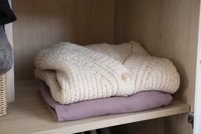 Une maille crème est pliée sur une tablette en bois près d’un haut lavande