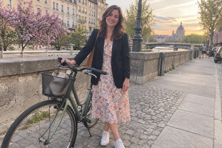 Une femme en blazer noir sur une robe midi fleurie et des baskets, à côté d’un vélo au bord de la Seine