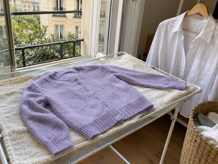 Un cardigan mauve posé à plat sur une serviette, à côté d’une chemise blanche sur cintre
