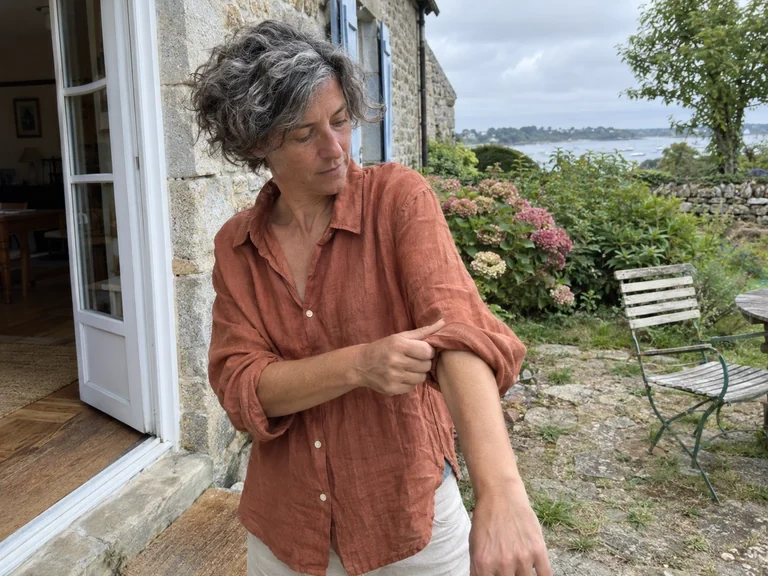 Une femme porte une chemise en lin terracotta devant une maison en pierre