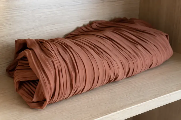 Un vêtement plissé terracotta est roulé souplement dans le sens des plis sur une étagère