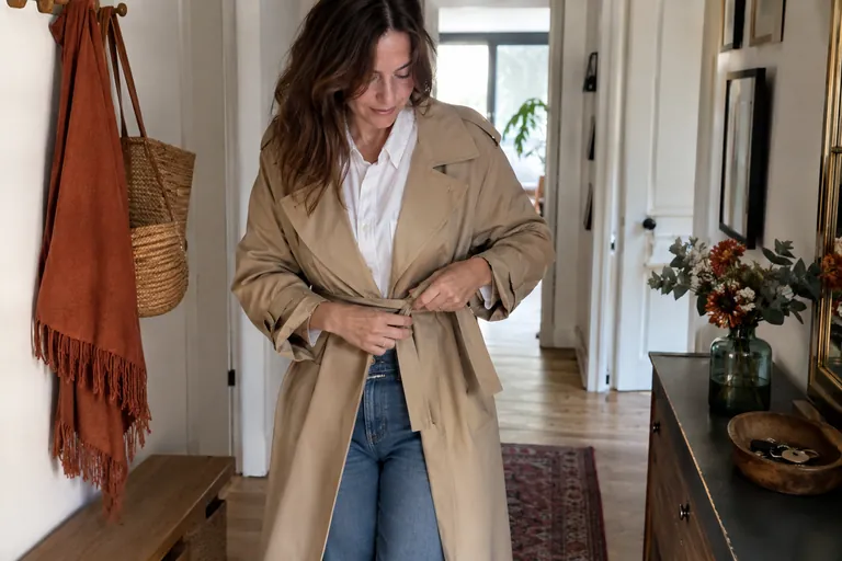 Une femme à la silhouette droite noue la ceinture d’un trench beige sur une chemise blanche