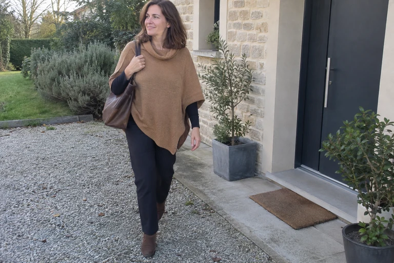 Mujer con un poncho camel sobre una blusa y un pantalón oscuros junto a una entrada.
