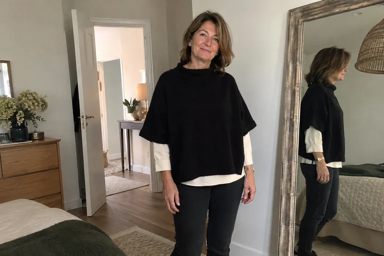 Woman wearing a short black poncho over a white shirt beside a mirror.
