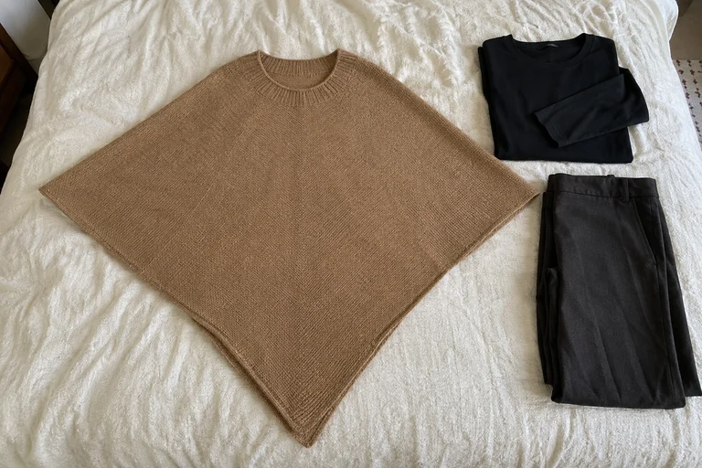 Diamond-shaped camel poncho laid flat beside a folded dark top and trousers.