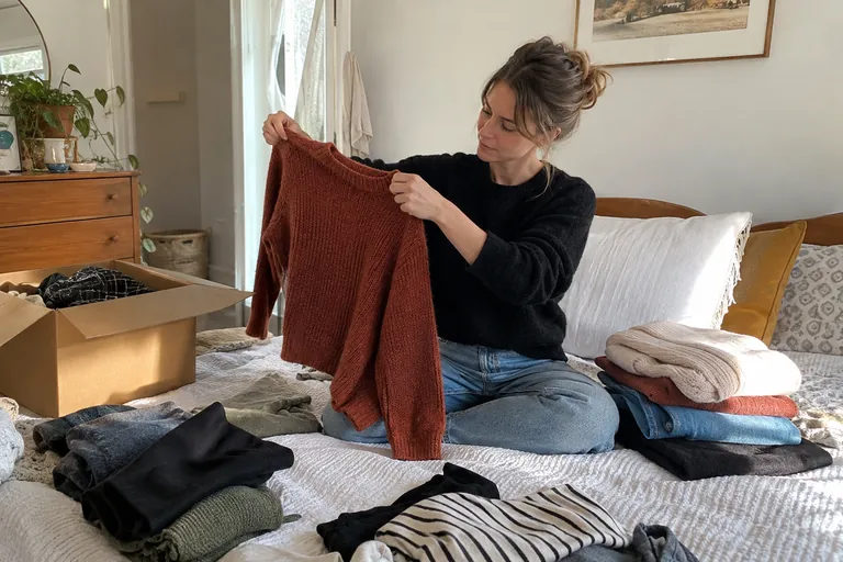 Une femme assise sur son lit trie ses vêtements en piles et examine un pull